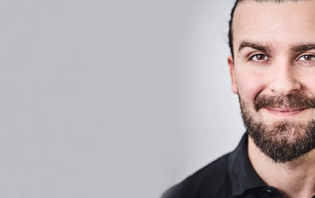 Porträt von MD Sarantis Adamakis, einem lächelnden Mann mit dunklem Bart und zu einem Zopf gebundenen dunklen Haaren vor einem neutralen, hellen Hintergrund.