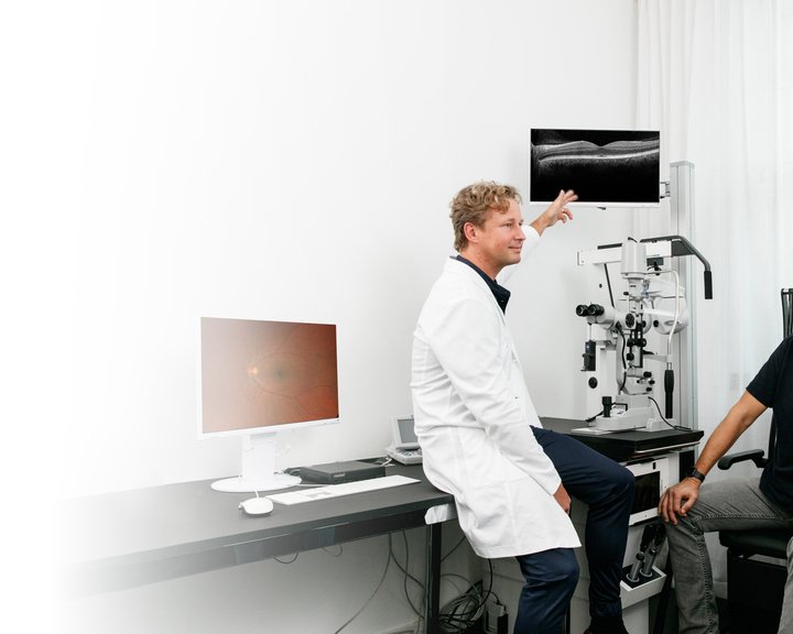 Ein Augenarzt in weißem Kittel erklärt einem Patienten die Ergebnisse einer OCT-Netzhautuntersuchung an einem Monitor in einem hellen Untersuchungsraum.