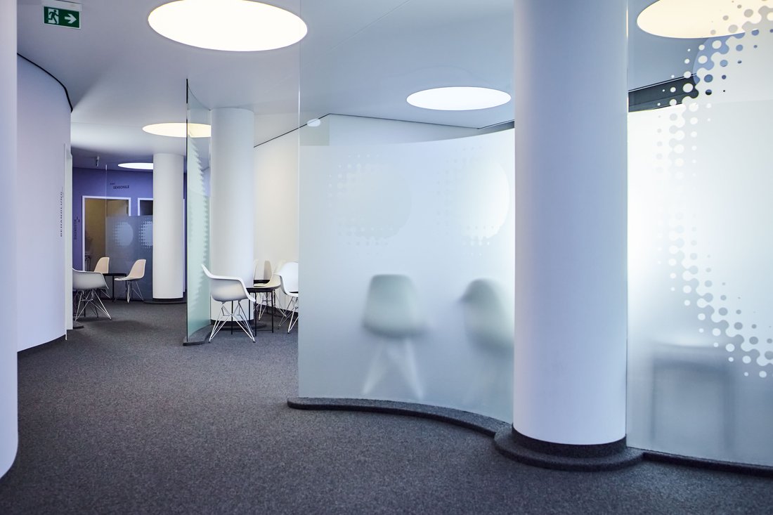Ein moderner, geschwungener Praxisflur mit runden Deckenleuchten, weißen Säulen und matten Glastrennwänden, hinter denen Sitzgelegenheiten für Patienten angeordnet sind.