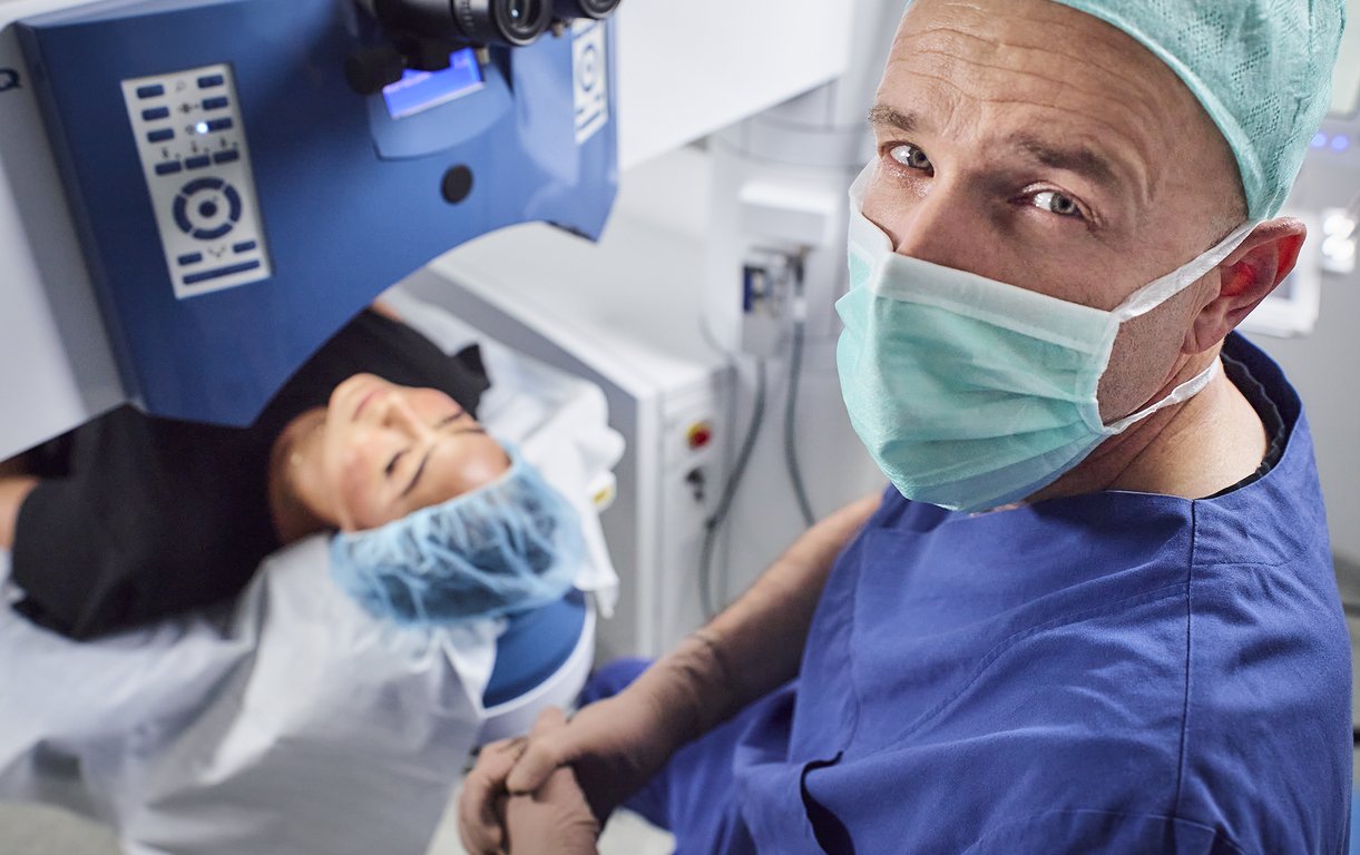 Ein Chirurg in blauer OP-Kleidung blickt in die Kamera, während eine Patientin unter einem Augenlasergerät für eine Behandlung positioniert ist.