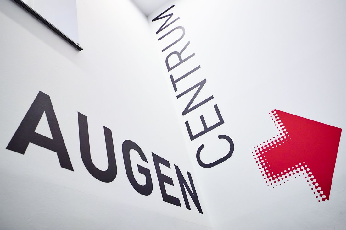 Eine Innenansicht zeigt eine weiße Wand an einer Treppe, auf der in großen, schwarzen Buchstaben die Wörter „AUGEN“ und „CENTRUM“ sowie ein markanter roter Richtungspfeil zur Orientierung für Patienten angebracht sind.