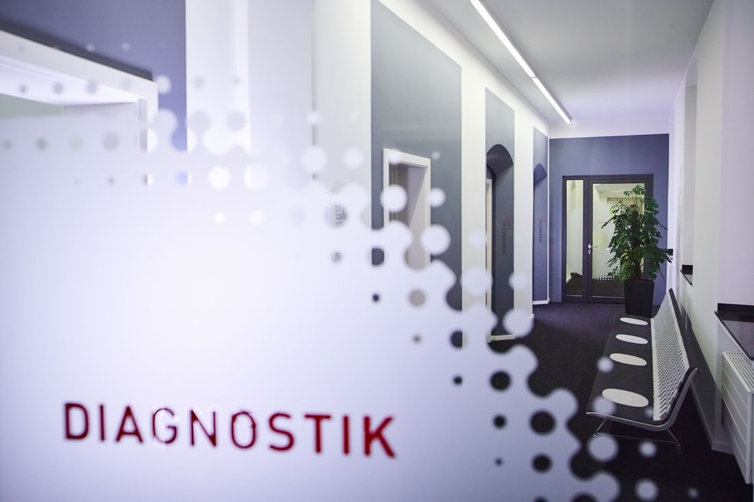 Blick durch eine Glastür mit der roten Aufschrift „DIAGNOSTIK“ in einen modern gestalteten, hellen Praxisflur mit einer langen Sitzbank und hohen Decken.