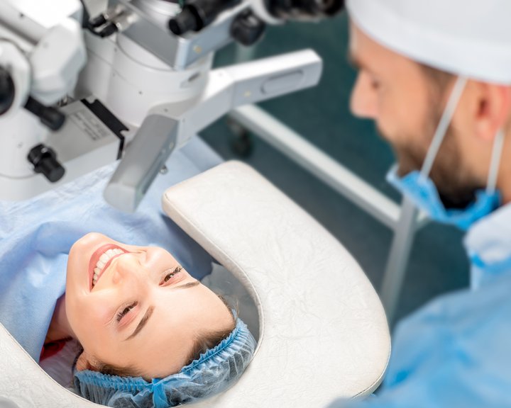 Eine lächelnde Patientin mit OP-Haube liegt unter einem chirurgischen Mikroskop bereit für eine Augenoperation, während ein Chirurg in blauer Schutzkleidung sie betreut.