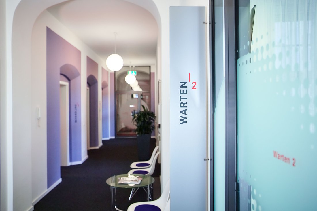 Ein heller, moderner Praxisflur mit hohen Decken und Rundbögen; an einer Glaswand im Vordergrund markiert ein Schild den Bereich „Warten 2“ neben einer Sitzgelegenheit.