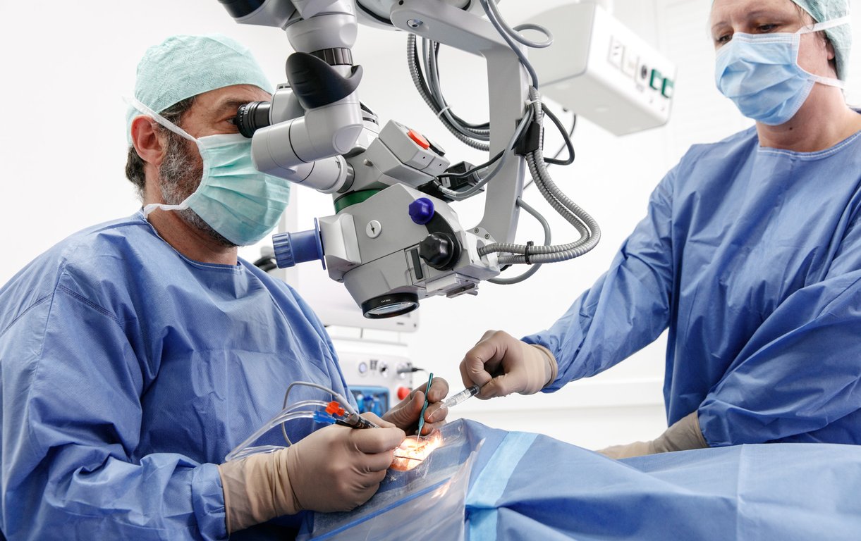 Ein Chirurg in blauer OP-Kleidung führt mithilfe eines großen Operationsmikroskops einen präzisen Eingriff am Auge eines Patienten durch, unterstützt von einer OP-Assistentin.