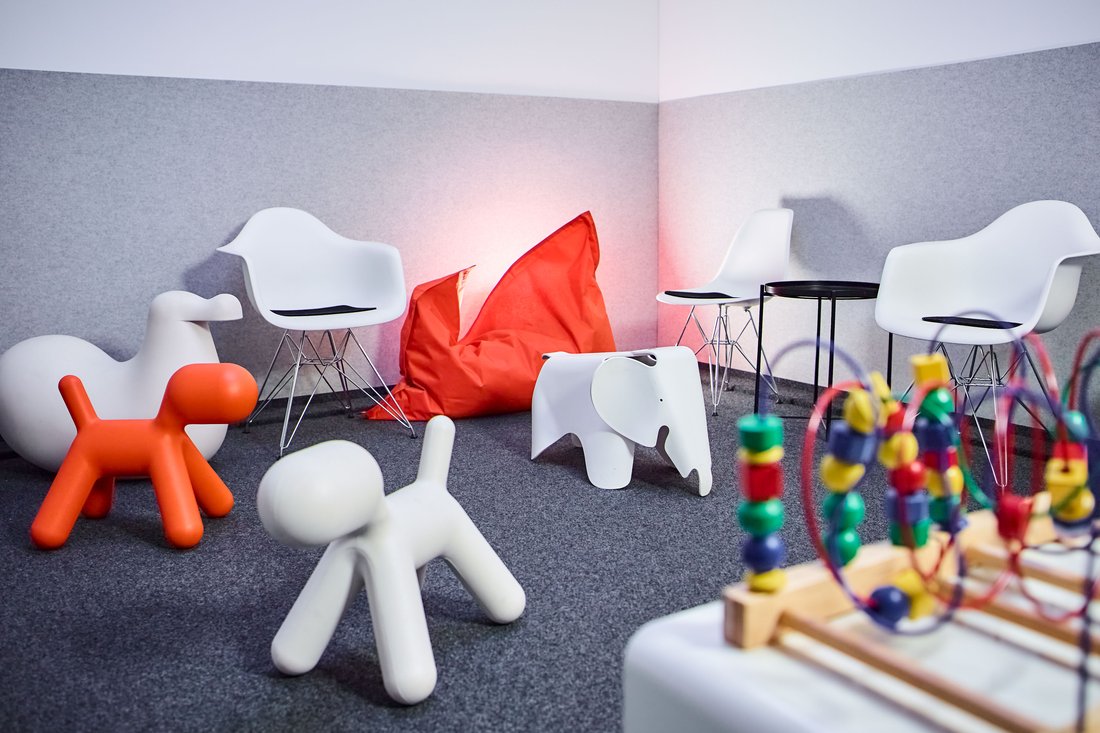 Ein farbenfroher Wartebereich für Kinder mit weißen Design-Sesseln, einem roten Sitzsack und verschiedenen Spielgeräten, darunter stilisierte Tierfiguren und ein Motorikschleifen-Spielzeug im Vordergrund.