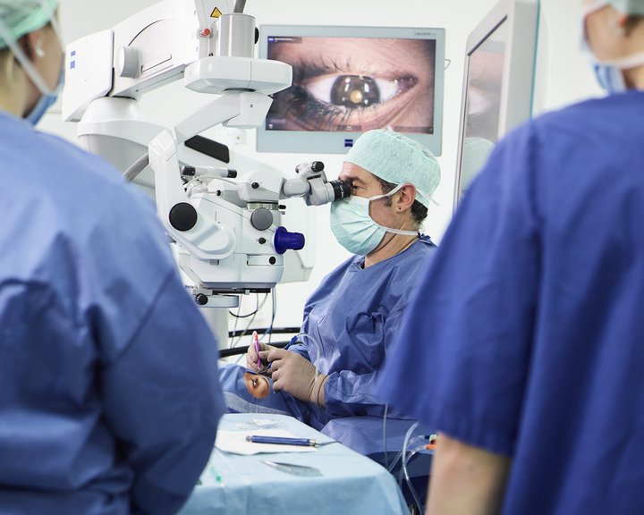 Ein Chirurg führt unter einem ZEISS-Operationsmikroskop eine mikrochirurgische Augenoperation durch, während das vergrößerte Auge auf einem Monitor im Hintergrund sichtbar ist.