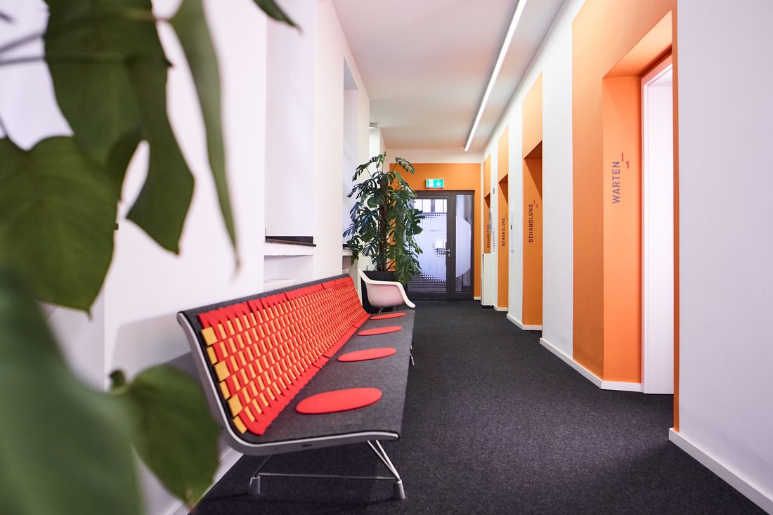 Ein moderner, heller Praxisflur mit orangefarbenen Wandakzenten; im Vordergrund steht eine stylische Sitzbank in Rot und Gelb, während im Hintergrund Schilder die Bereiche „Warten 1“ und „Behandlung“ kennzeichnen.