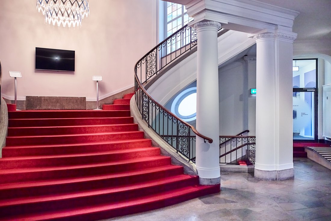 Ein repräsentativer Eingangsbereich mit einer breiten, rot teppichbelegten Treppe, die von klassizistischen weißen Säulen und einem kunstvollen schmiedeeisernen Geländer gesäumt wird.