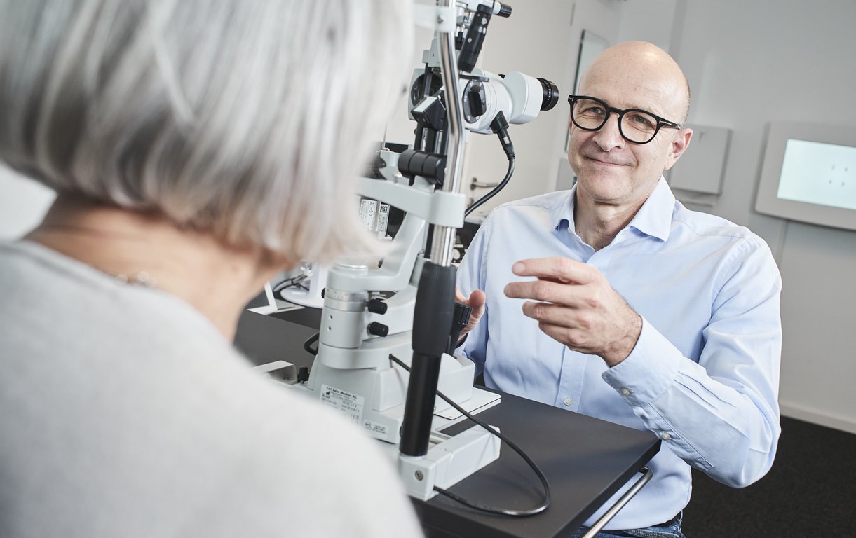 Ein lächelnder Augenarzt mit Brille sitzt einer Patientin mit hellem Haar gegenüber und bereitet eine Untersuchung an einer Spaltlampe vor.