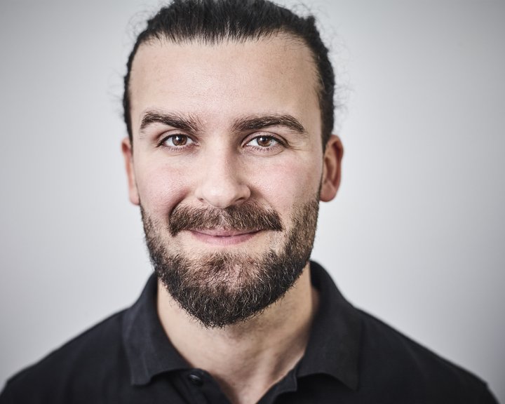Porträt von MD Sarantis Adamakis, einem lächelnden Mann mit dunklem Bart und zu einem Zopf gebundenen dunklen Haaren vor einem neutralen, hellen Hintergrund.