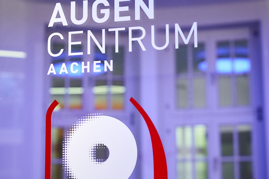 Das Logo des „AUGEN CENTRUM AACHEN“ ist in weißer Schrift auf einer Glasfläche angebracht, ergänzt durch ein abstraktes rotes Augen-Symbol, während im Hintergrund klassische Fenster und blaues Deckenlicht sichtbar sind.