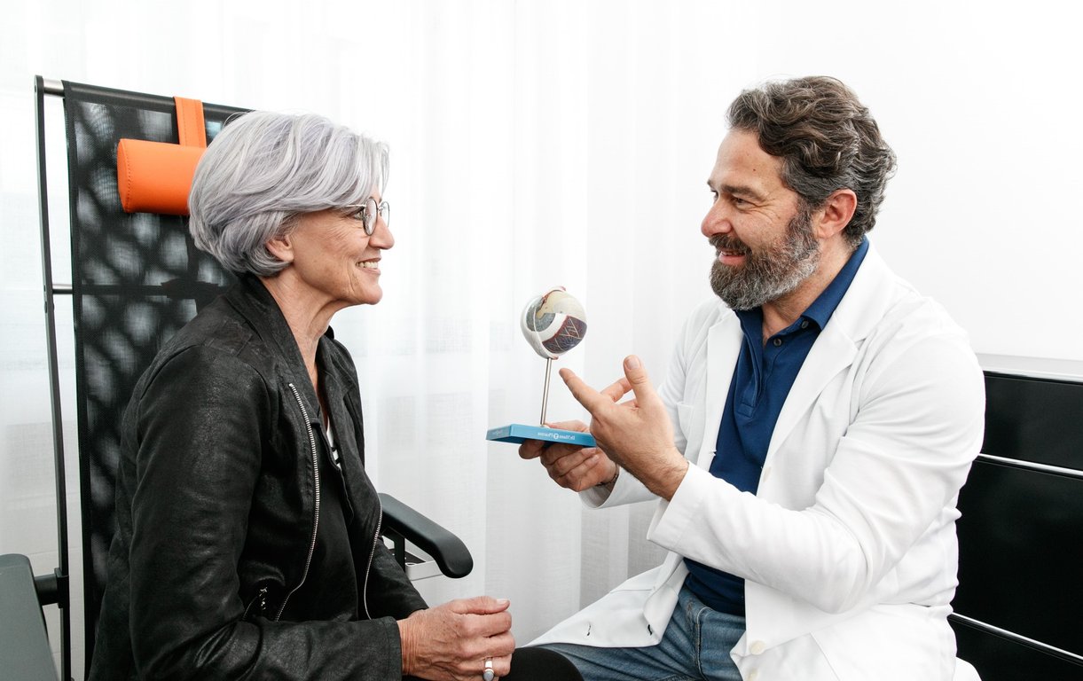 Ein bärtiger Augenarzt im weißen Kittel erklärt einer Patientin mit grauem Haar einen Befund, indem er auf ein kleines, dreidimensionales anatomisches Modell des menschlichen Augapfels zeigt.