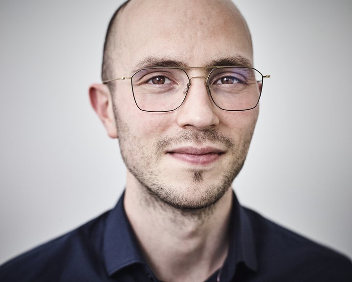 Porträt von Dr. med. Joel Mor, einem lächelnden Mann mit Glatze und einer feinen, dunklen Brille vor einem neutralen, hellen Hintergrund.